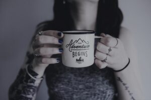 Tattooed hands holding an enamel mug with the inscription "The adventure begins"