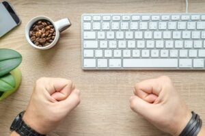 A desk with a keyboard, cup full of coffee beans and a person's clenched fists resting on the desk