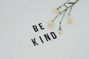 The words 'be kind' on a white background with a spray of white carnation flowers beside them