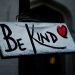 A rectangular wooden board tied to a post outside a building with the words Be Kind and a red heart painted on the board's white background