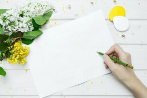 A blank piece of white paper with a person's hand hovering holding a green crayon, with white and yellow flowers beside the paper on a white table top