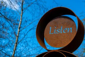 Photograph of a sculpture with a metal disc set inside a circular tube with the word listen cut out, all set against a blue sky