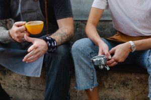 Two anonymous co-workers sitting outside talking, one holding a coffee cup and the other the stump filter of a coffee machine.